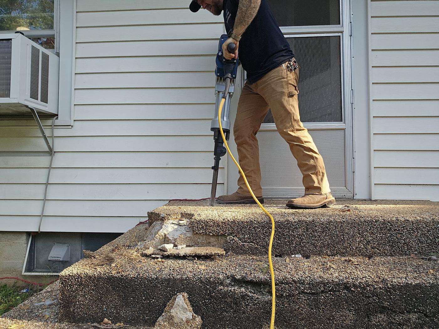 A demolition hammer breaking out an old concrete stoop, the worker in a Man Handy Solutions shirt