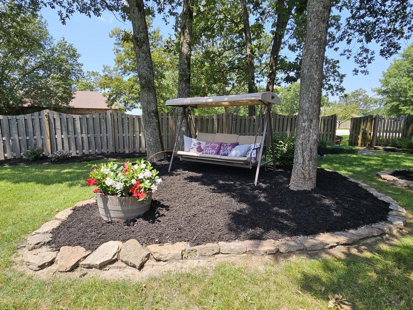 A stone-edged mulch bed under mature oaks with a porch swing and a whiskey-barrel planter of red and white flowers
